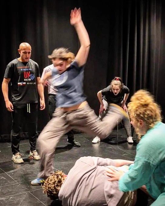 An example of stage combat in a performance. 
Image of a person laid on the floor with people looking on as someone is about to kick them in the head.