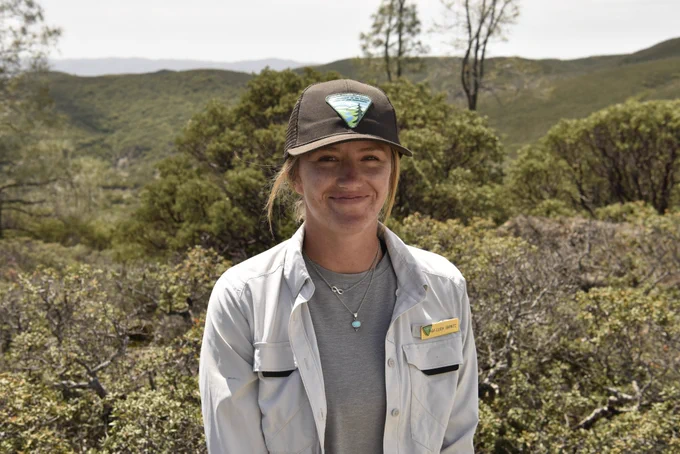A woman wearing a dark baseball cap, light button-up shirt, and gray t-shirt stands outdoors in front of a backdrop of green, forested hills. 