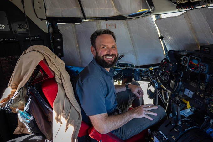  A man with a beard, wearing a short-sleeved gray shirt, sits smiling in the cockpit of a large airtanker. 