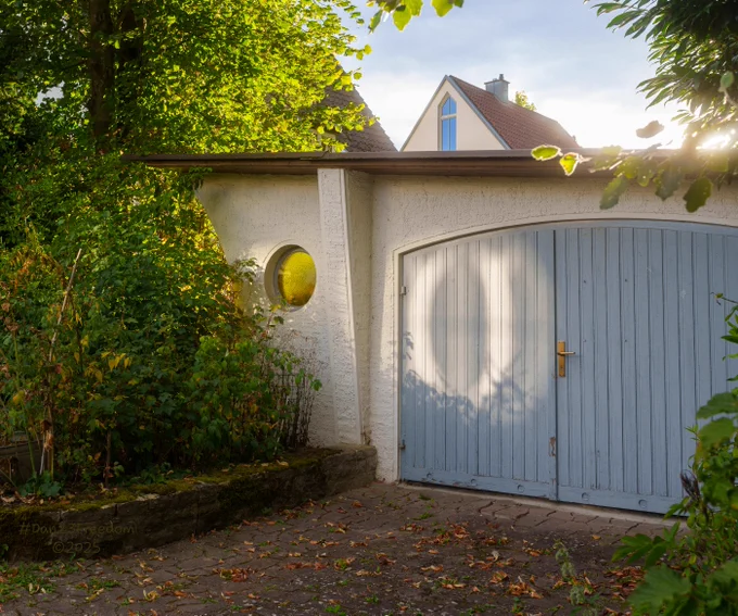 A captivating, sun-drenched image features a light blue wooden garage door and a textured white stucco wall, framed by lush green foliage. A large diffuse reflection glows brightly on the garage door, its source a delightful mystery. To the left, a small, circular window with yellow-orange, textured glass shines like a warm, watchful eye. The scene is bathed in golden hour light, creating vibrant highlights on the leaves and subtle shadows that add depth to the textured surfaces. Strong natural framing with a f/8 aperture lends excellent depth of field, keeping both the intimate details and the broader context in sharp focus. — The golden hour, a mischievous painter, cast an ethereal heart upon the garage door, while the little round window, an ancient eye, watched the street turn to dusk.