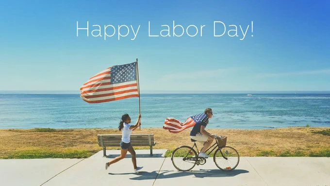 Happy Labor Day greeting above an image of two individuals, one waving an American flag and the other riding a bicycle, by the seaside under a clear blue sky.