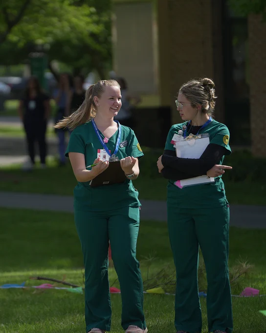 Two FCC nursing students stand together outside. They are wearing green scrubs and holding clipboards.