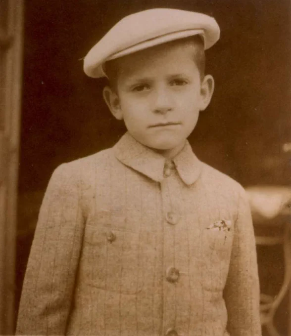 Vintage sepia-tone photo of a young boy in a double-breasted coat and a flat cap.

