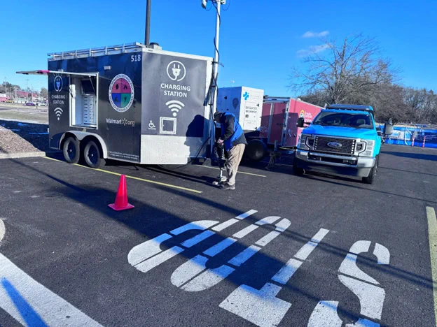 A person connects an electric vehicle to a mobile charging station trailer in a parking lot under clear blue skies.