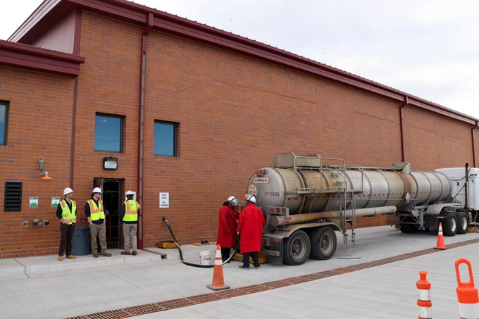 Three Highlands Ranch Water employees wearing yellow safety vests and white hardhats standing next to a large brick building overlooking a chemical delivery that two men in long red coats and hardhats are handling next to a large tanker truck.