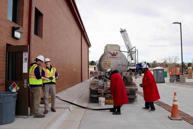 Two Highlands Ranch Water employees wearing yellow safety vests and hardhats, discussing plans while two men wearing long red safety coats and white hardhats prepare chemical load from a tanker truck at a large chemical building with cloudy skies in the background.