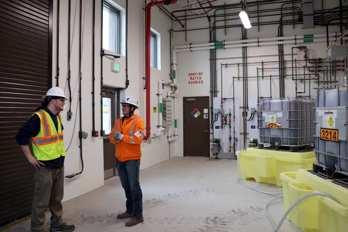 Two men talking in a water treatment chemical storage room, one is wearing a yellow safety vest and hardhat and another is wearing an orange safety jacket and hardhat.