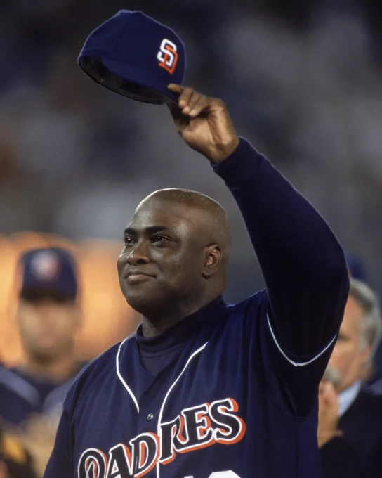Tony Gwynn holds his blue Padres hat in the air with his left hand and salutes the crowd. He is wearing a blue Padres uniform as well.