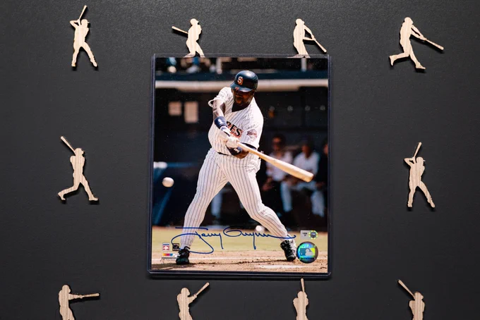A signed photo of Tony Gwynn sits in the middle of a black background with wooden figures swinging a bat. Tony Gwynn is wearing a white Padres jersey with a dark blue helmet and orange and white lettering.