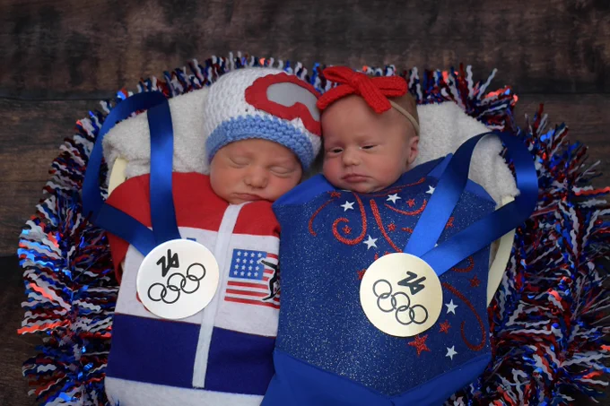Two sleeping babies in a basket are dressed in patriotic outfits with medals. One wears a white and red knit hat and an outfit with a USA flag. The other wears a red bow and a starry blue outfit. The medals say '24 and Olympic rings'.
