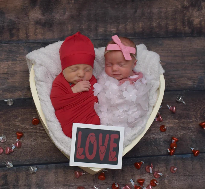Two sleeping infants, wrapped in red and flower-patterned white cloths respectively, lie in a heart-shaped basket lined with white fabric. A sign reads \"LOVE\" with heart shapes in the letters. Hershey's kisses surround the basket on the wood surface.