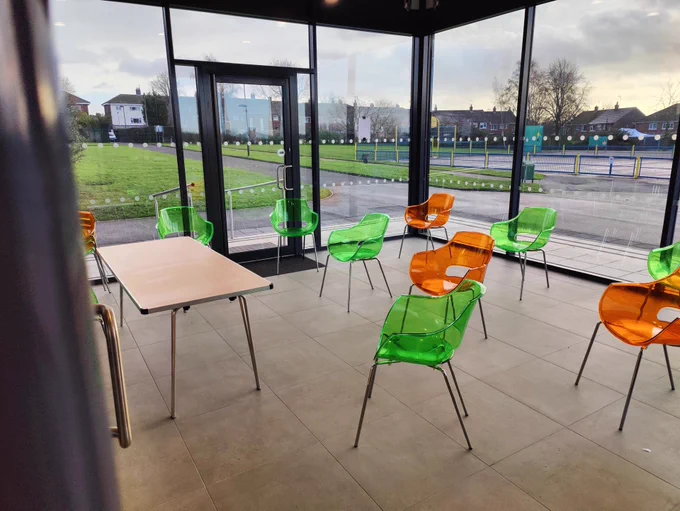 Empty room with transparent green and orange chairs arranged around a light wooden table near large glass windows overlooking a grassy area.