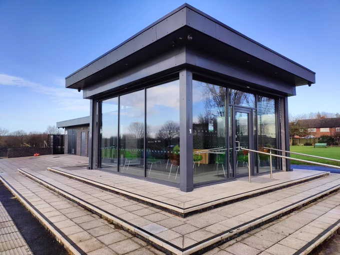Modern glass pavilion with metal framing on a tiled platform overlooking a green field under a clear sky.