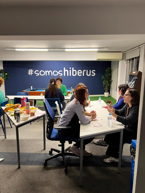 Grupo de mujeres sentadas en parejas en varias mesas, con una mesa de snacks en el centro. Están charlando y riendo entre ellas. En la pared de color azul del fondo se lee #somosHiberus