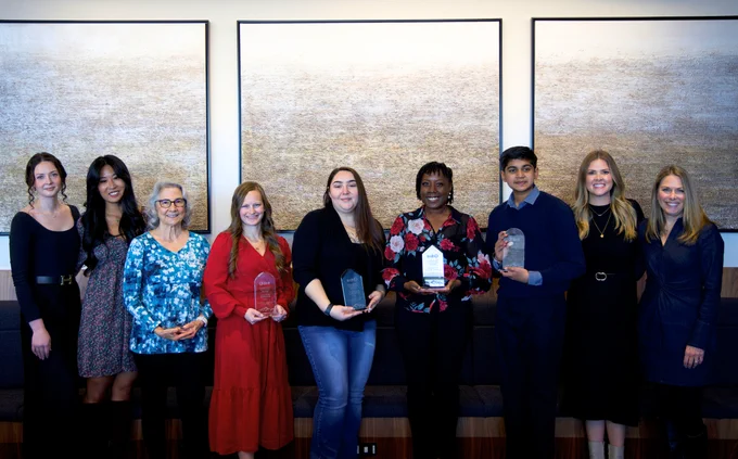 A group of nine people standing in front of three large abstract paintings. Five individuals in the center hold glass awards. They are in a well-lit room with a bench in the background.
