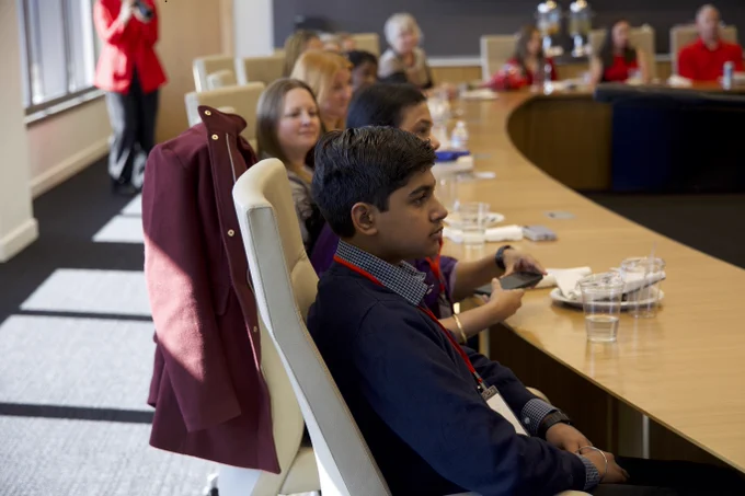 A group of people seated around a large, curved conference table in a well-lit office. A person in a red jacket is standing near the window. Cups and papers are on the table.