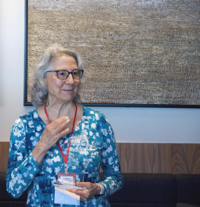 A person with gray hair and glasses is standing in front of a textured artwork on a wall. They are holding a napkin and gesturing with one hand. The person is wearing a blue patterned shirt and a red lanyard.