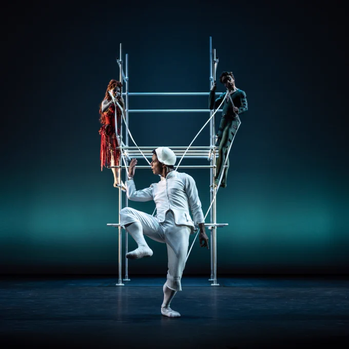 Marcelino Sambé in a white costume poses dynamically in the foreground, balancing on one leg. In the background, Matthew Ball and Mayara Magri stand on a metal scaffold structure, one dressed in red and the other in black. The stage is dimly lit with a blue gradient backdrop.