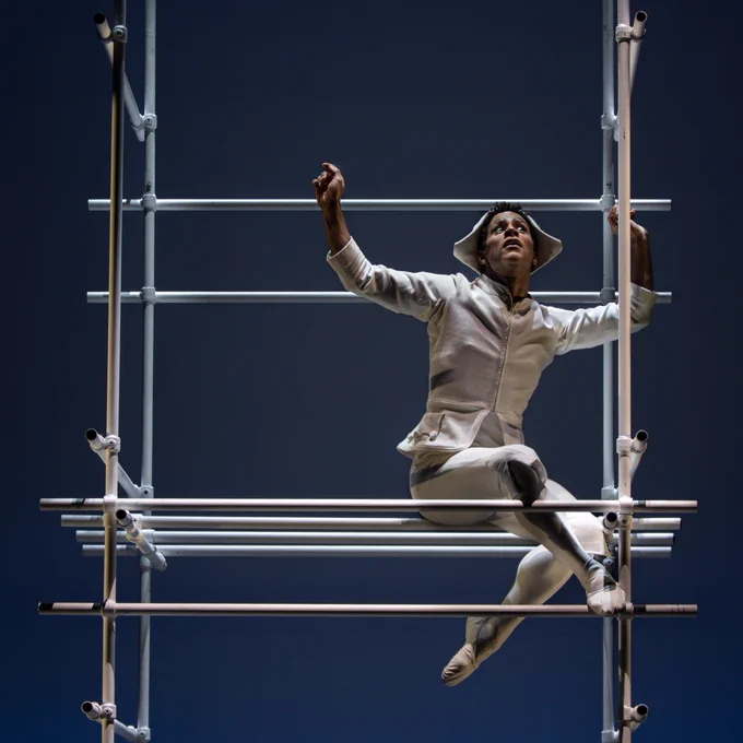 Marcelino Sambé  in a white costume and hat is poised elegantly on a metal scaffolding structure against a dark blue background.