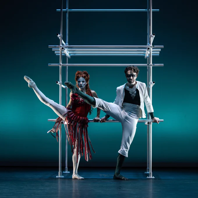 Matthew Ball and Mayara Magri perform on stage. Mayara Magri, on the left, is in a red and black costume, while Matthew Ball, on the right, wears a white suit with dramatic makeup. They are in front of a structure made of horizontal and vertical bars, with a blue-green background.