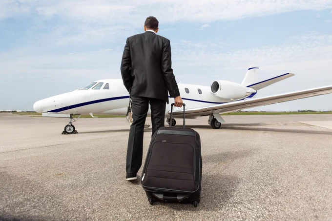 Person in a business suit walking towards a private jet while pulling a suitcase on a tarmac.