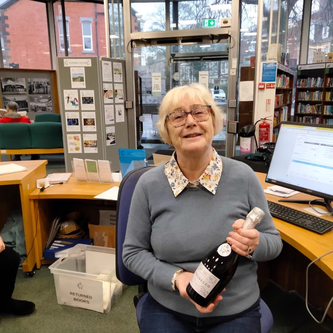 Image of Stephanie, sitting at the Library desk, holding bottle of Prosecco
