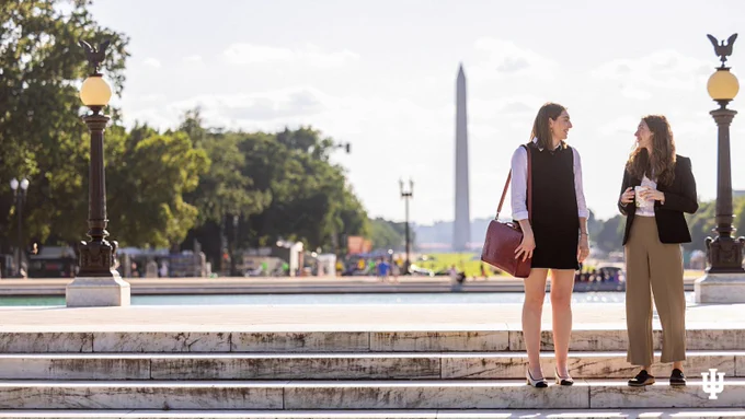 Two people are standing on a set of marble steps, engaged in conversation. In the background, the Washington Monument is visible, surrounded by trees and open space. The scene is under a clear sky with a few clouds.
