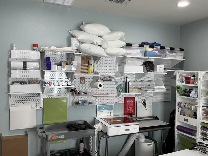 White metal pegboard wall organization system above a craft workstation, holding shelves with pillows, storage bins, paper supplies, tools, scissors, and rolls of material, with a cutting machine and cart below in a clean, organized creative workspace.
