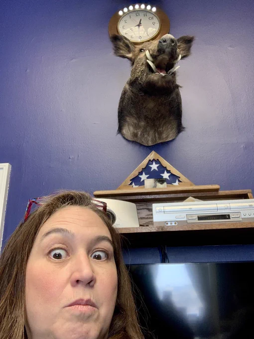 A woman making a surprised face near a stuffed boar on a wall and a clock