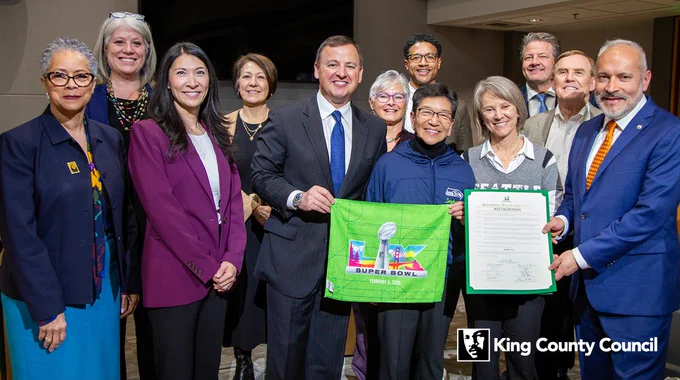 Mary Yu with councilmembers and others. A Seahawks Super Bowl LX towel and the recognition document are held in the foreground.