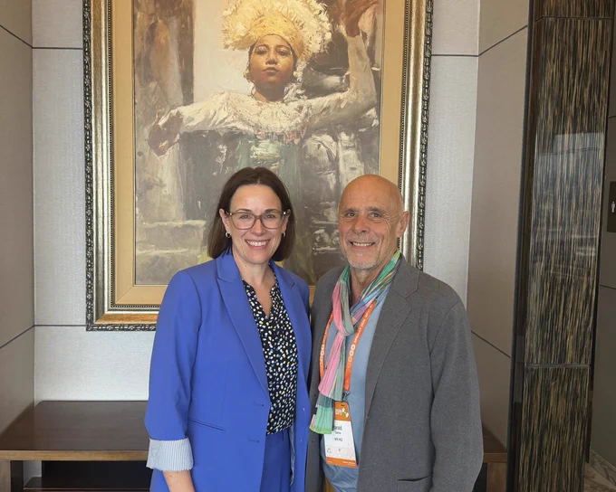 Two people smile and pose together in front of a painting. 