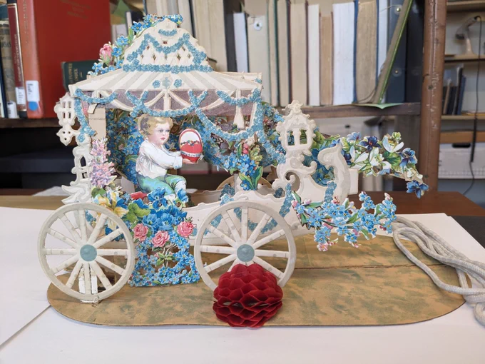 on a white surface, a white carriage covered in blue flowers with a small child inside the carriage holding a red heart that says "my valentine."