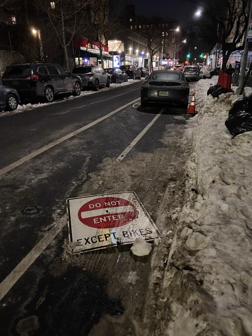A “Do not enter, except bikes” sign lies in slush and grime in a parking lane in between two cars parked in a bike lane, while beyond and behind additional cars are parked in the bike lane. A wall of snow and ice partially blocks the parking lane.