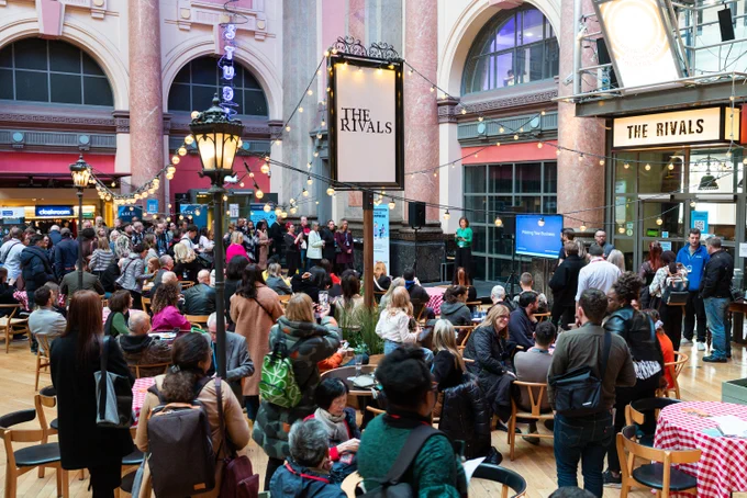 Crowded indoor event at The Rivals with people seated and standing, engaged in conversations under string lights and arched windows.