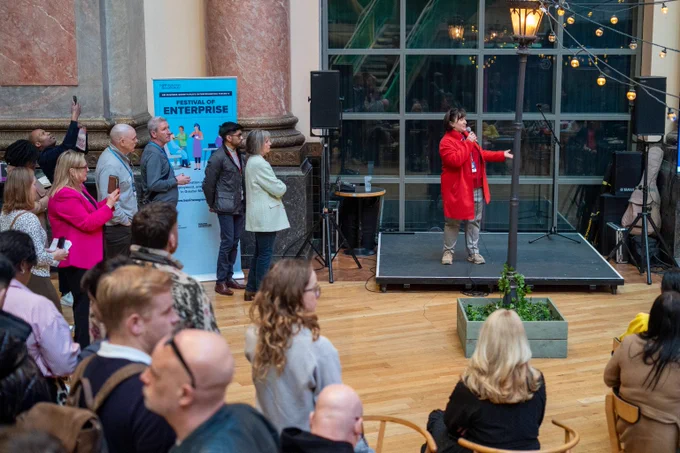 A woman in a red coat speaks on stage at a Festival of Enterprise event with an attentive audience in a spacious indoor venue.