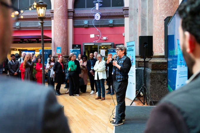Man speaking to an attentive crowd in a spacious indoor venue with marble columns and wooden floors.