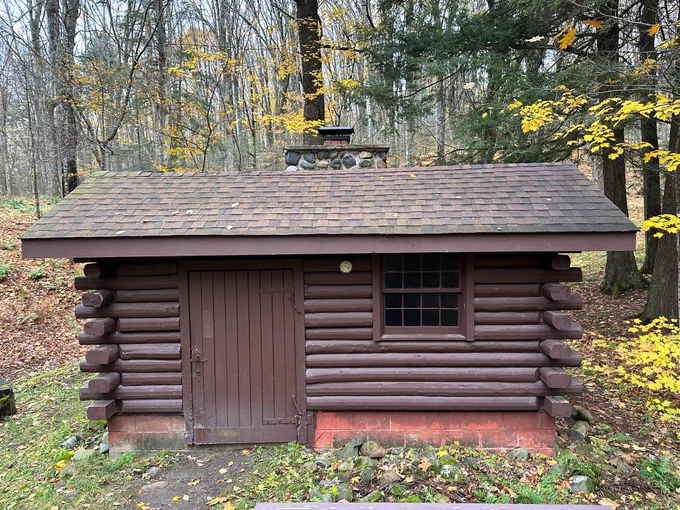 A small log warming shack with a shingled roof sits in a wooded area, surrounded by trees and fallen leaves, with a stone chimney visible behind the building. 
