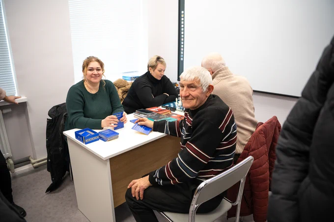 Four residents of Zavodske, including two women and two men, sit around a table inside the newly rehabilitated community hub, engaging in conversation and activities in a bright, restored space designed for essential services and community gatherings.