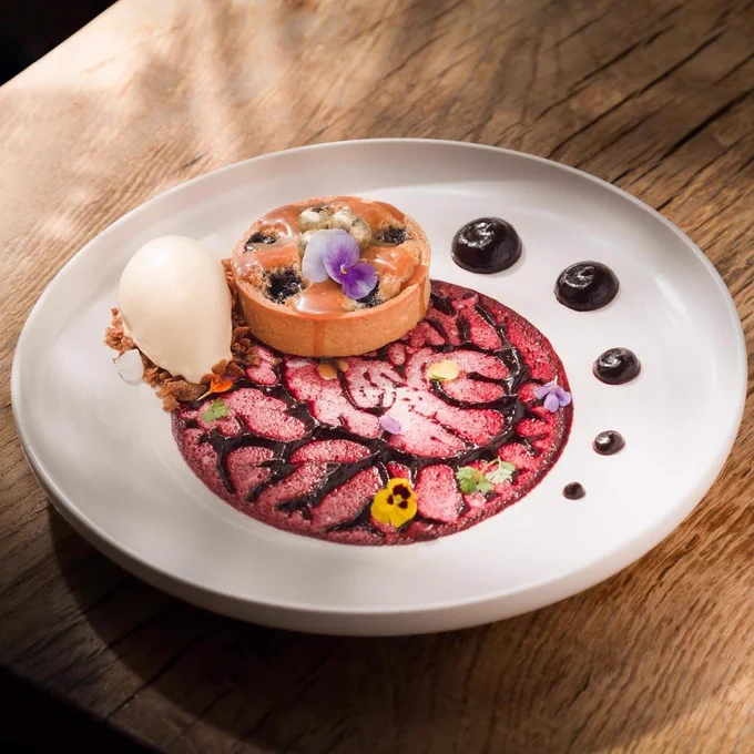 Round tart with edible flowers and cream, served with a scoop of white sorbet and artistic berry sauce on a white plate.