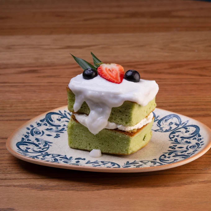 Two-layer green tea sponge cake topped with white cream, a strawberry slice, blueberries, and mint leaves on a decorative plate.