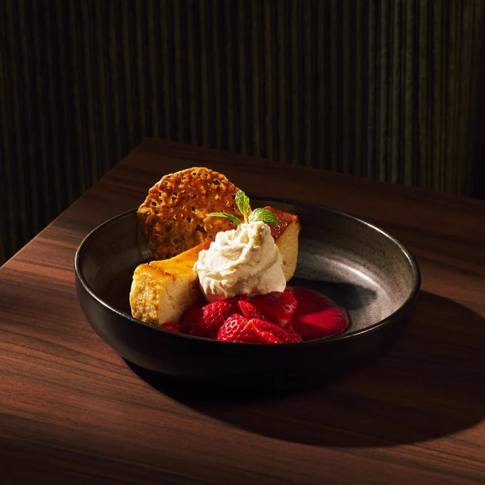 Slice of cheesecake topped with whipped cream and a mint leaf, served with fresh strawberries and a crisp cookie in a black bowl.