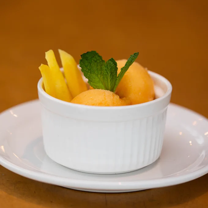 Two scoops of orange sorbet garnished with fresh mint leaves in a white ramekin on a white plate.