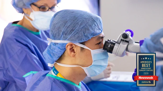 A healthcare professional in surgical attire is looking through a microscope, while another person in similar attire assists nearby. The area is equipped for a medical procedure. A plaque in the foreground reads "America's Best Ambulatory Surgery Centers 2026" from Newsweek.