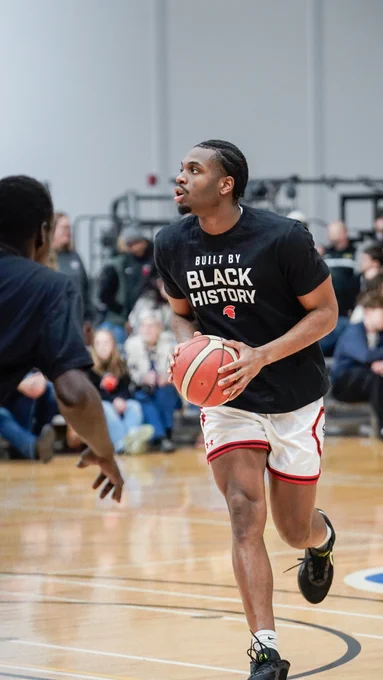 A basketball player in motion on an indoor court, wearing a shirt that reads "Built By Black History," holds a basketball. Another player is nearby. Spectators are seated in the background.