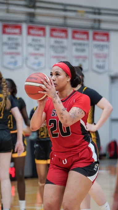 A basketball player wearing a red jersey with the number 20 is focused and preparing to make a shot. In the background, several players in black and yellow jerseys are visible. Banners hang on the wall in the gymnasium.