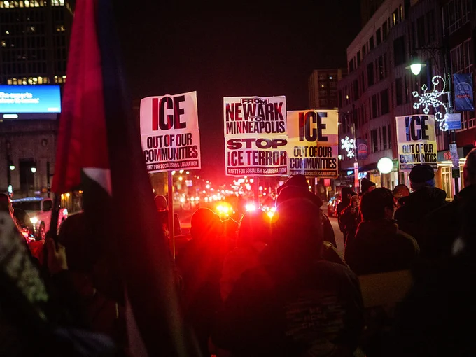 a group of people hold signs that say from newark to minneapolis end ice terror at night and red police lights flash 