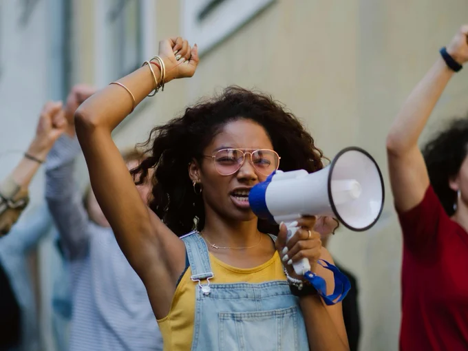 A young woman wearing dungarees over a yellow t-shirt walks with a group of protesters. She raises one hand above her head and she holds a megaphone on the other.