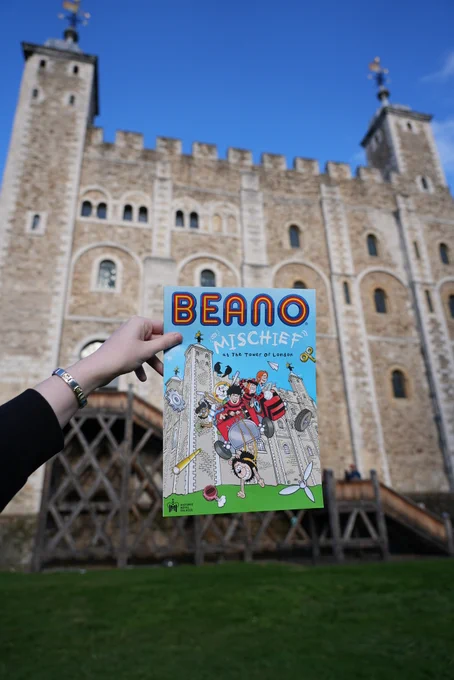 A person holds a Beano comic pamphlet against the backdrop of the White Tower at the Tower of London on a sunny day.