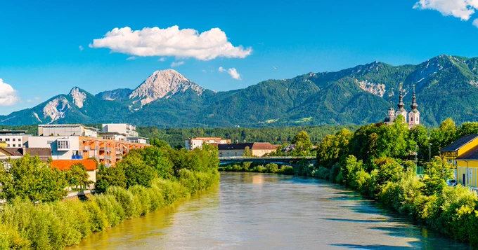 A picturesque view of a river flowing through a vibrant town, surrounded by lush greenery. In the background, the mountainous landscape includes Mittagskogel, under a clear blue sky with a few clouds. A church with tall spires is visible on the right, and a bridge crosses the river.
