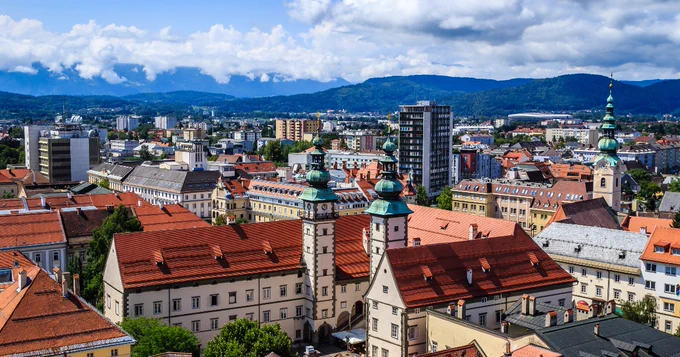 A panoramic view of Ljubljana, Slovenia, featuring a mix of historical and modern architecture. Prominent are buildings with red-tiled roofs and ornate spires, set against a backdrop of lush hills and cloudy skies.